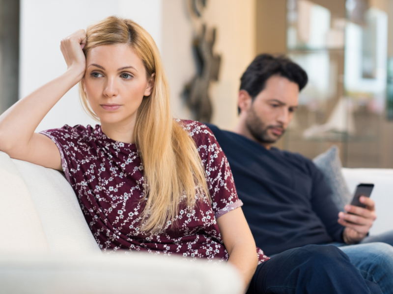 Unhappy woman sits on couch while man uses phone