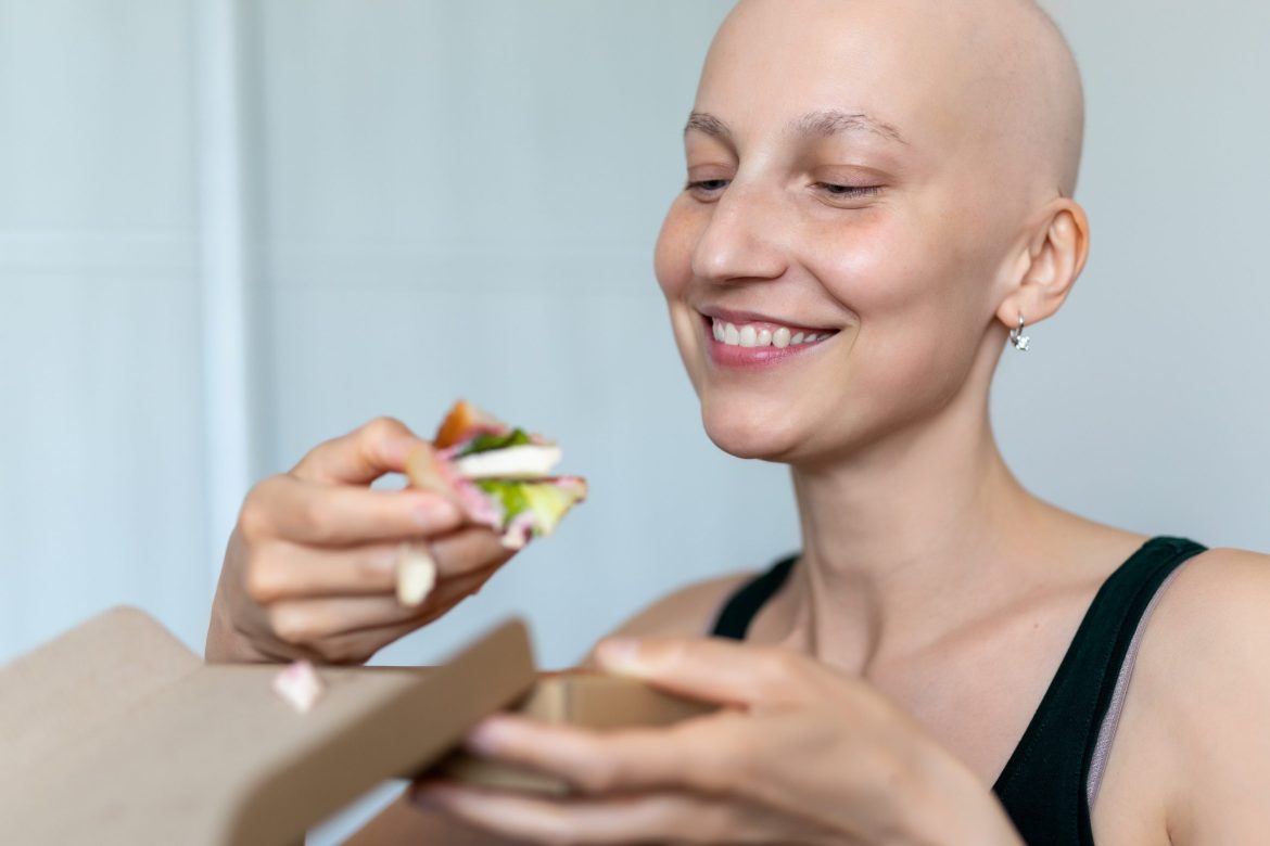 woman with bald head due to cancer smiling and eating a sandwich