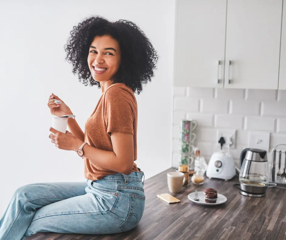 A smiling woman sitting on a kitchen counter, enjoying a cup of probiotic yogurt in a modern setting