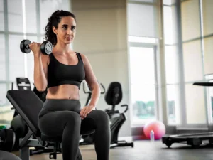 A fit woman in black activewear performing a bicep curl with a dumbbell while sitting on a bench in a well-lit modern gym