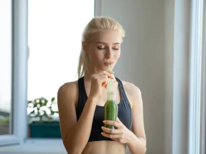 A fit woman in athletic wear sipping a green juice through a straw, standing by a bright window