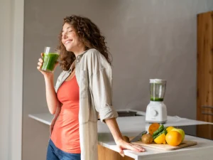 A cheerful woman in a kitchen enjoying a green smoothie, with fresh fruits and a blender on the counter