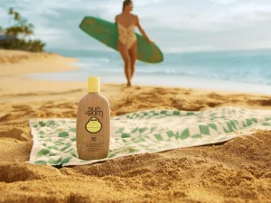 Sun Bum sunscreen bottle on sandy beach with surfer nearby